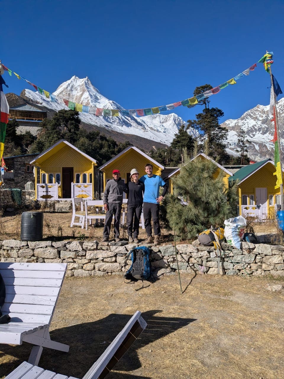 Three trekkers by yellow lodges, snow-capped Manaslu, and prayer flags