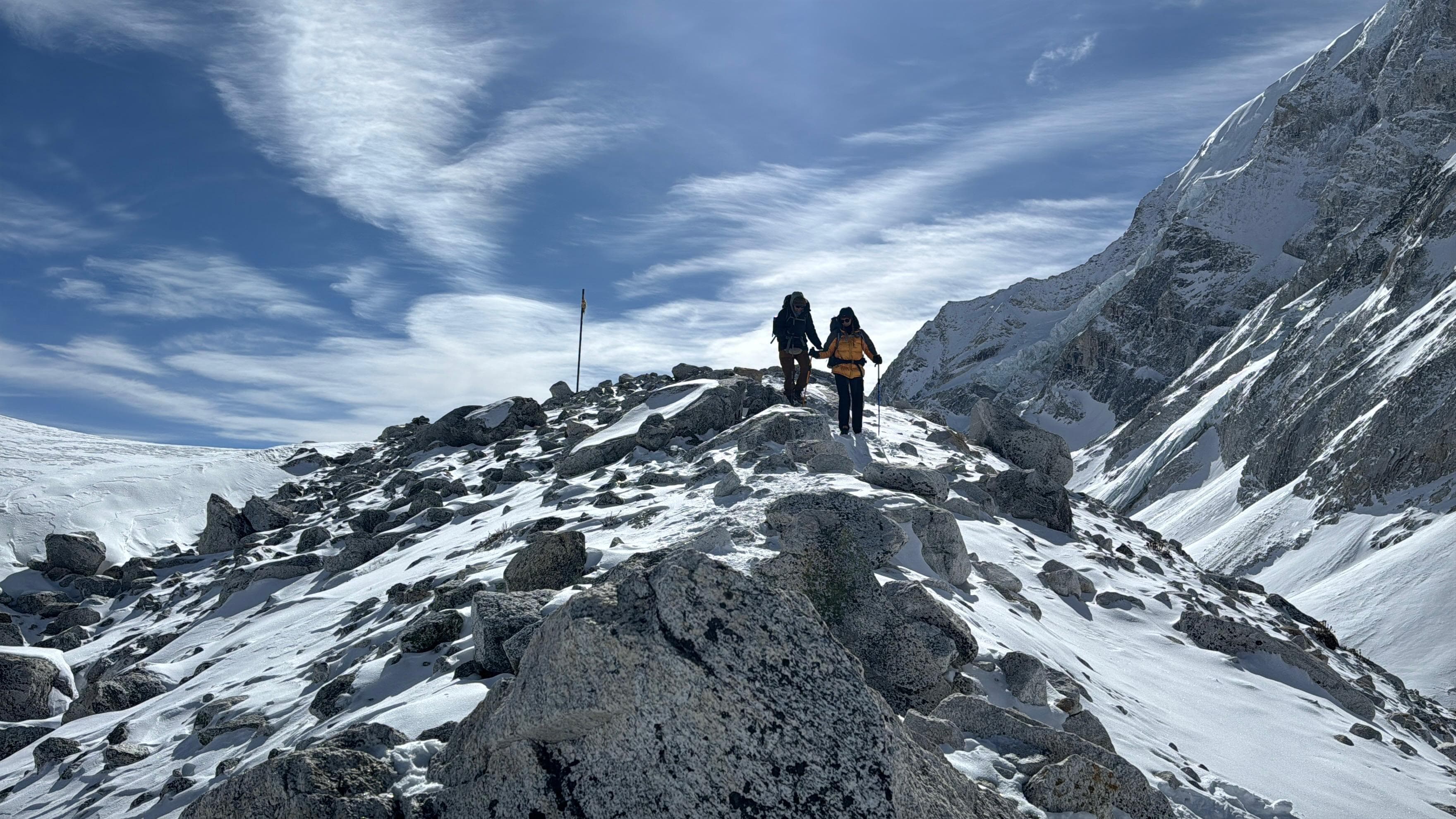 Two trekkers on a snowy, rocky Himalayan trail under blue sky