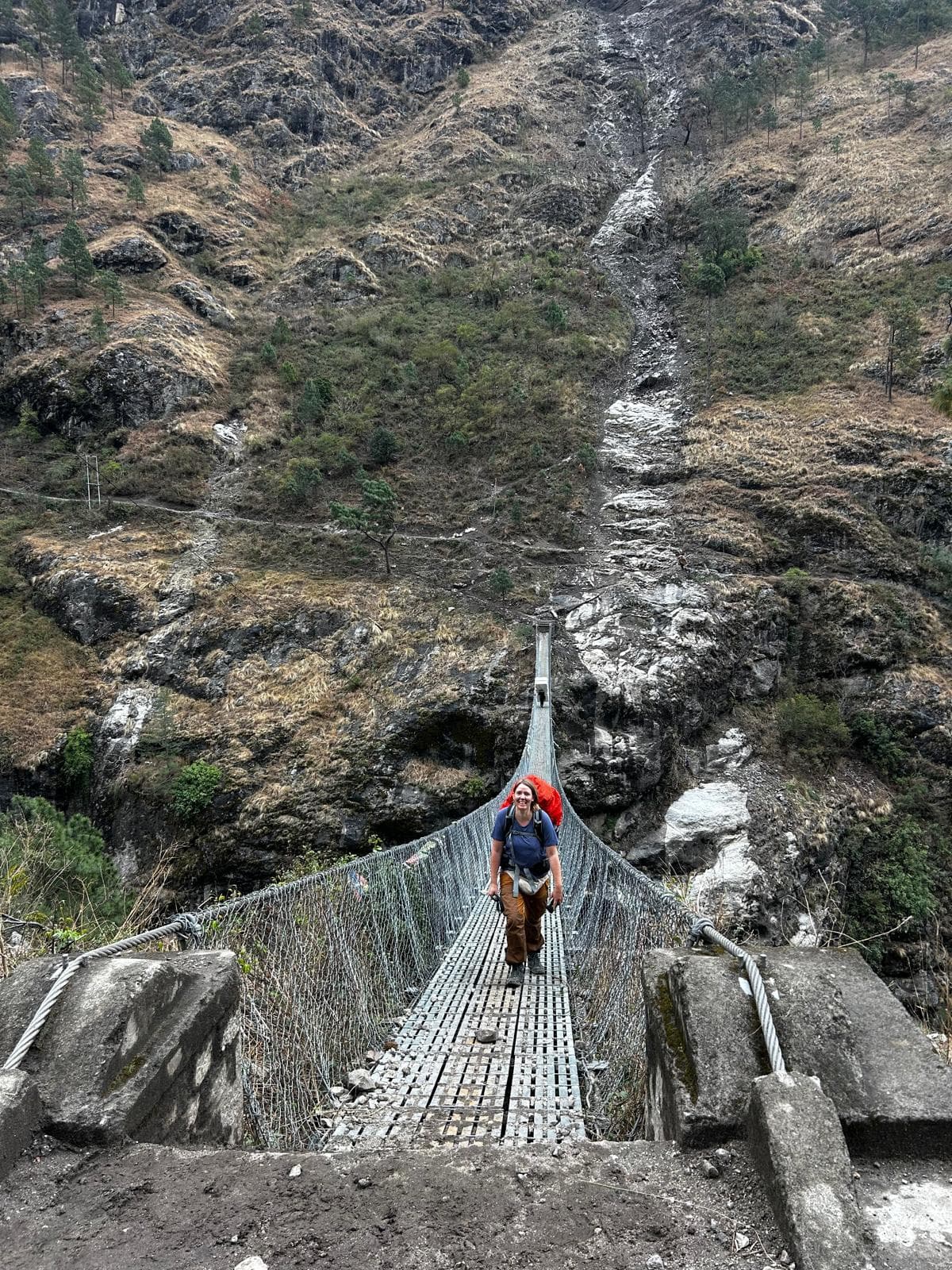 Smiling trekker crosses metal suspension bridge amidst rugged Nepal mountains