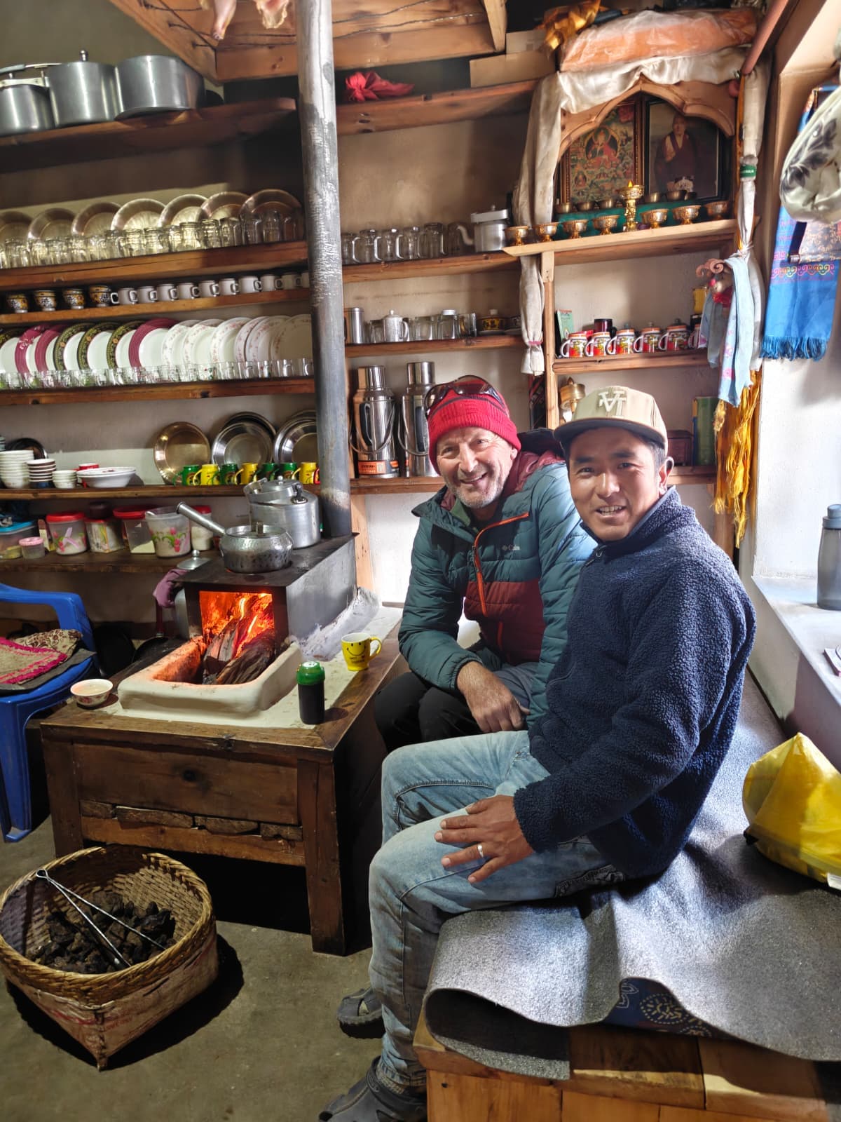 Two smiling men in a rustic Himalayan teahouse beside a wood-burning stove